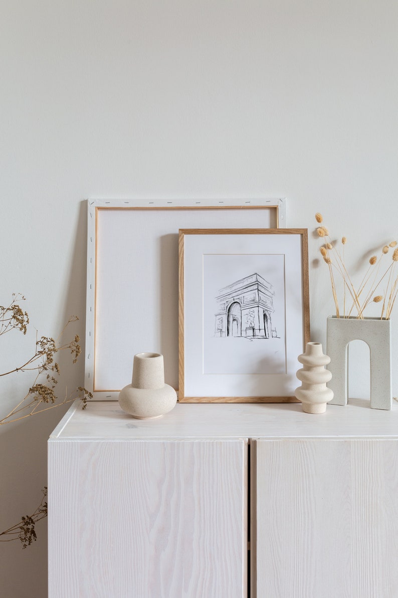 May include: A white cabinet with two doors, a framed black and white drawing of the Arc de Triomphe, a white vase, a beige vase, and dried flowers in a gray vase.