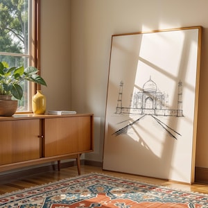 May include: A framed black and white line drawing of the Taj Mahal on a beige background. The frame is made of natural wood and has a light brown finish. The drawing is centered on the canvas and is surrounded by a white border. The drawing is set against a beige wall and a wooden floor. A wooden cabinet with two doors is in the foreground. A green plant is in a brown pot on the windowsill.