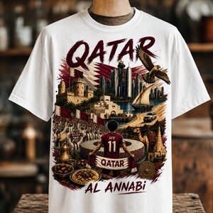 May include: White t-shirt featuring a detailed graphic of Qatar's skyline, a falcon, and a soccer player. The word "QATAR" is prominently displayed in a burgundy font. The shirt also includes the text "AL ANNABI".