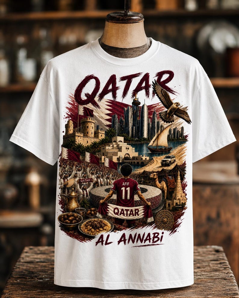 May include: White t-shirt featuring a detailed graphic of Qatar's skyline, a falcon, and a soccer player. The word "QATAR" is prominently displayed in a burgundy font. The shirt also includes the text "AL ANNABI".
