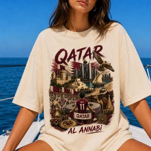 May include: Cream-colored t-shirt with a graphic design featuring the word "QATAR" in burgundy, a cityscape, a falcon, and a soccer player. The shirt also includes the text "AL ANNABI".