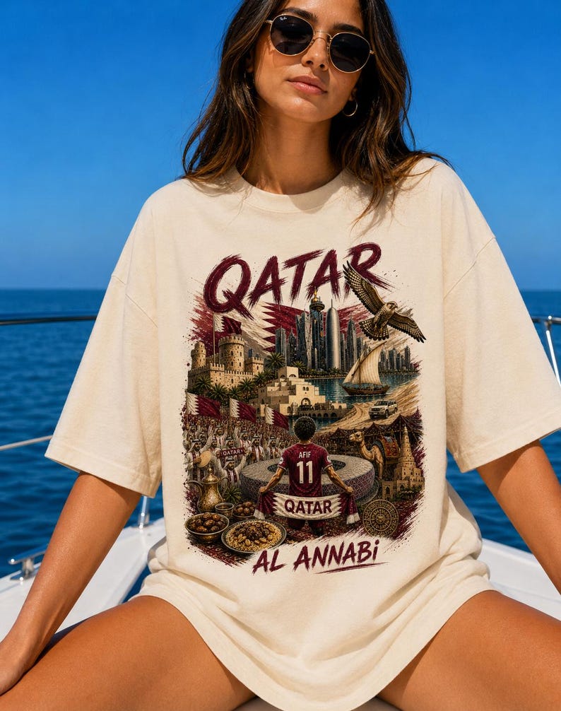 May include: Cream-colored t-shirt with a graphic design featuring the word "QATAR" in burgundy, a cityscape, a falcon, and a soccer player. The shirt also includes the text "AL ANNABI".