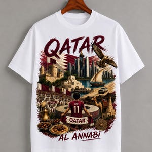 May include: White t-shirt featuring a detailed graphic of Qatar with the text "QATAR" in maroon. The design includes a cityscape, a soccer player, and the words "AL ANNABI".