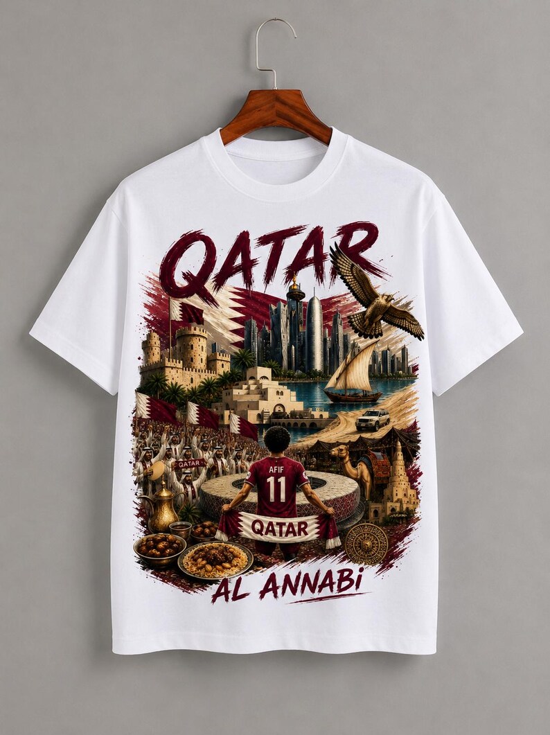 May include: White t-shirt featuring a detailed graphic of Qatar with the text "QATAR" in maroon. The design includes a cityscape, a soccer player, and the words "AL ANNABI".