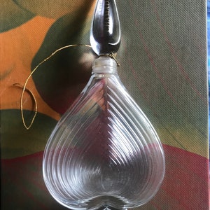 Könnte beinhalten: Eine durchsichtige Glasparfümflasche in Herzform mit einer strukturierten Oberfläche. Die Flasche hat einen durchsichtigen Glasverschluss mit dem Wort "Chamade" darauf graviert. Die Flasche steht auf einer strukturierten Oberfläche.