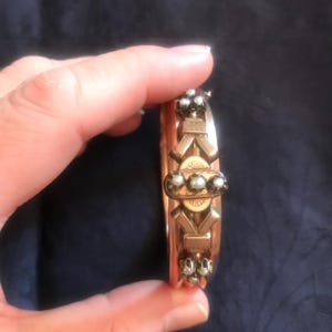 Op de afbeelding: Een goudkleurige scharnierende armband met een centrale rechthoekige plaat versierd met drie kleine witte parels. Het metalen oppervlak vertoont gedetailleerde gravures, waaronder strikachtige motieven en extra clusters van parels.