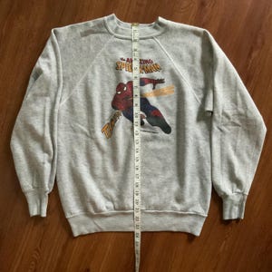 K&ouml;nnte beinhalten: Graues Sweatshirt mit Rundhalsausschnitt und Spider-Man-Grafik. Das Design enth&auml;lt den Text "The Amazing Spider-Man" und "Thwip!". Das Sweatshirt ist etwa 61 cm lang. Das Kleidungsst&uuml;ck liegt flach auf einer Holzoberfl&auml;che.