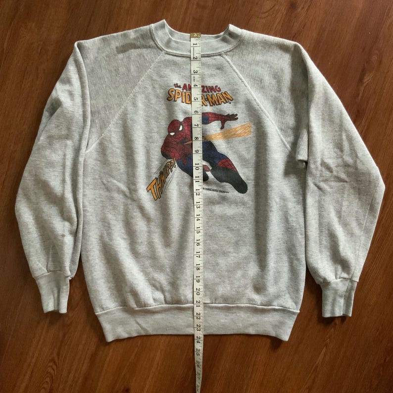 K&ouml;nnte beinhalten: Graues Sweatshirt mit Rundhalsausschnitt und Spider-Man-Grafik. Das Design enth&auml;lt den Text "The Amazing Spider-Man" und "Thwip!". Das Sweatshirt ist etwa 61 cm lang. Das Kleidungsst&uuml;ck liegt flach auf einer Holzoberfl&auml;che.