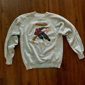 K&ouml;nnte beinhalten: Hellgraues Sweatshirt mit einem Aufdruck von Spider-Man. Der Superheld ist in seinem klassischen rot-blauen Anzug dargestellt, mit dem Schriftzug "THE AMAZING SPIDER-MAN" dar&uuml;ber. Das Sweatshirt hat lange &Auml;rmel und einen Rundhalsausschnitt.