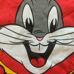 May include: Close-up of a Bugs Bunny graphic on a red background. The cartoon rabbit has gray fur, large white eyes, and a wide, smiling mouth. The image is from a Looney Tunes design.