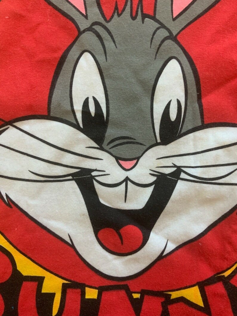 May include: Close-up of a Bugs Bunny graphic on a red background. The cartoon rabbit has gray fur, large white eyes, and a wide, smiling mouth. The image is from a Looney Tunes design.