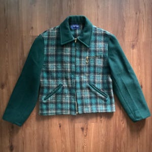 Pode incluir: Casaco vintage com padrão xadrez em verde-azulado, castanho e preto. As mangas e a gola são verde-azulado liso. O casaco tem fecho de correr e dois bolsos frontais. A etiqueta do casaco diz "SPORTCLAS".