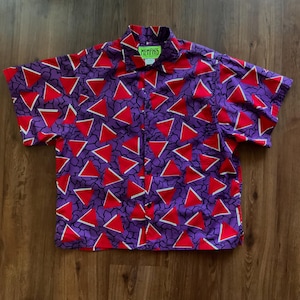 May include: A short-sleeved button-down shirt with a vibrant geometric pattern. The shirt features a purple background with red triangles and white accents. The collar has a green tag that reads "MEMPHIS". The shirt is laid flat on a wooden surface.