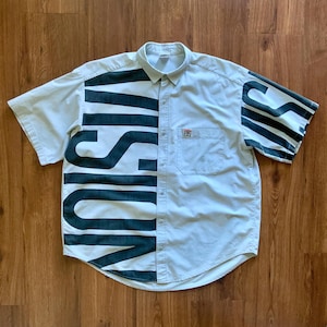 May include: A short-sleeved button-down shirt in a light beige color. The word "VISION" is printed in large, dark green letters across the front. The sleeves have dark green stripes. A small pocket is on the left chest.