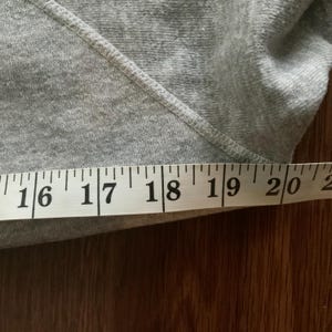 K&ouml;nnte beinhalten: Nahaufnahme eines grauen Sweatshirts mit einem Ma&szlig;band. Das Band zeigt ein Ma&szlig; von etwa 40,6 bis 50,8 cm. Das Sweatshirt hat eine diagonale Naht &uuml;ber das Bild.