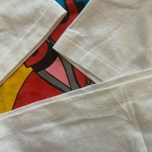 May include: Close-up of a white long-sleeved shirt with a colorful graphic design. The graphic features red, blue, yellow, and pink elements. The sleeves are folded, showing the seams and fabric texture.