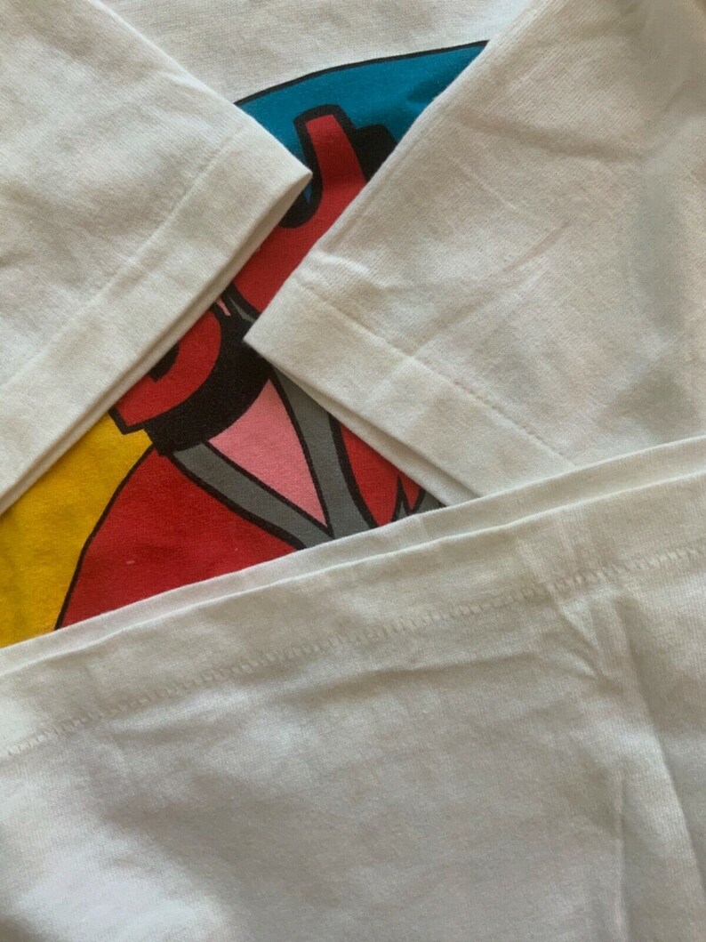 May include: Close-up of a white long-sleeved shirt with a colorful graphic design. The graphic features red, blue, yellow, and pink elements. The sleeves are folded, showing the seams and fabric texture.