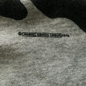 K&ouml;nnte beinhalten: Nahaufnahme eines grauen Stoffes mit einem schwarzen Design. Der Text "MARVEL COMICS GROUP 1979" ist schwarz gedruckt. Der Stoff hat ein strukturiertes Aussehen.