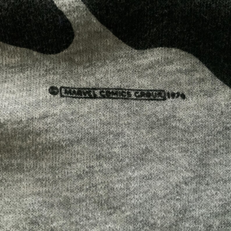 K&ouml;nnte beinhalten: Nahaufnahme eines grauen Stoffes mit einem schwarzen Design. Der Text "MARVEL COMICS GROUP 1979" ist schwarz gedruckt. Der Stoff hat ein strukturiertes Aussehen.