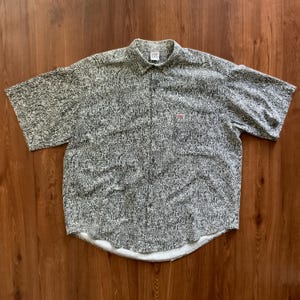 Puede incluir: Camisa de manga corta con botones y un estampado abstracto en blanco y negro. La camisa tiene un pequeño logotipo rojo en el bolsillo del pecho y un dobladillo blanco. El cuello tiene una pequeña etiqueta.