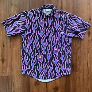 May include: A short-sleeved button-down shirt with a purple and black flame pattern. The shirt has a small pocket on the left side and features a cartoon character design.