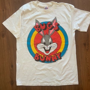 May include: White Bugs Bunny t-shirt featuring a Bugs Bunny graphic in a red, yellow, and blue circle. The words "BUGS" and "BUNNY" are in red letters. The shirt is made of a soft, comfortable material.