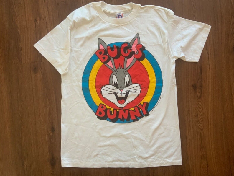 May include: White Bugs Bunny t-shirt featuring a Bugs Bunny graphic in a red, yellow, and blue circle. The words "BUGS" and "BUNNY" are in red letters. The shirt is made of a soft, comfortable material.