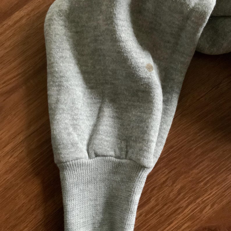 K&ouml;nnte beinhalten: Nahaufnahme eines grauen Sweatshirt-&Auml;rmels. Der &Auml;rmel hat ein geripptes B&uuml;ndchen und einen kleinen Fleck. Der Stoff scheint ein weiches, gestricktes Material zu sein. Der Hintergrund ist eine Holzoberfl&auml;che.
