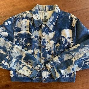 May include: A blue denim jacket with a white print of faces all over it. The jacket has a button closure and two pockets on the front. The label on the jacket says "The Hand."