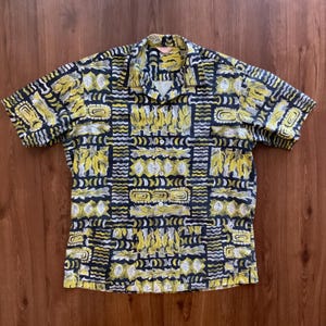 May include: Short-sleeved shirt with a bold, repeating pattern in yellow, black, and white. The design features geometric shapes and leaf-like motifs. The shirt has a classic collar and a relaxed fit, suitable for casual wear.