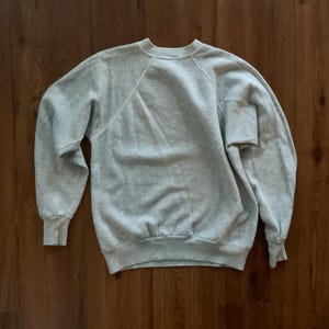 K&ouml;nnte beinhalten: Ein hellgraues Sweatshirt mit Rundhalsausschnitt und langen &Auml;rmeln. Das Sweatshirt hat ein Raglan&auml;rmel-Design und gerippte B&uuml;ndchen und Saum. Der Stoff scheint ein weiches, bequemes Material zu sein. Das Sweatshirt liegt flach auf einer Holzoberfl&auml;che.