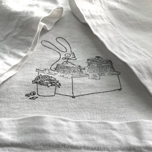 Op de afbeelding: Een wit T-shirt met een zwart-witte illustratie van een konijn dat aan een bureau zit met een stapel papier. Het konijn draagt een bril en heeft een verbaasde uitdrukking op zijn gezicht.