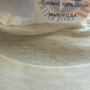 May include: Close-up of a white cotton t-shirt collar with a tag. The tag reads "Or Iron If Decorated, Do Not Bleach, Content: 100% Cotton, Made in U.S.A." The t-shirt is made of a soft, knitted fabric.