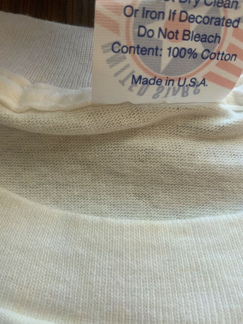 May include: Close-up of a white cotton t-shirt collar with a tag. The tag reads "Or Iron If Decorated, Do Not Bleach, Content: 100% Cotton, Made in U.S.A." The t-shirt is made of a soft, knitted fabric.