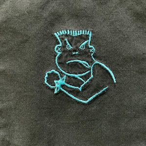 May include: Embroidered design of a cartoon character with a blue outline on a black fabric background. The character has a grumpy expression and is depicted with arms crossed.