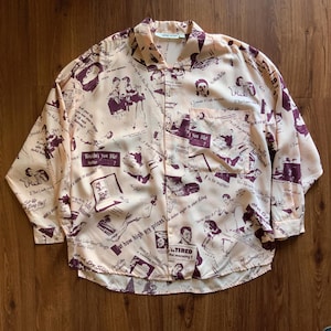 May include: A button-down shirt with a cream-colored background and a repeating pattern of illustrations and text in dark purple. The shirt has a pocket on the chest and long sleeves. The text includes phrases like "Wouldn't you like some?" and "Tired the morning?"