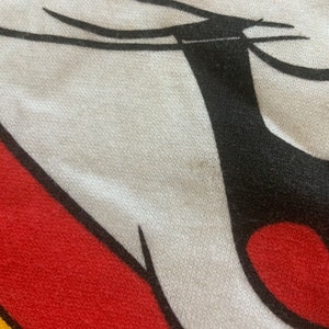May include: Close-up of a white, red, black, and yellow graphic design. The design features a cartoon character's face, with a white face, black eyes and ears, and a red mouth. The background is a textured fabric.