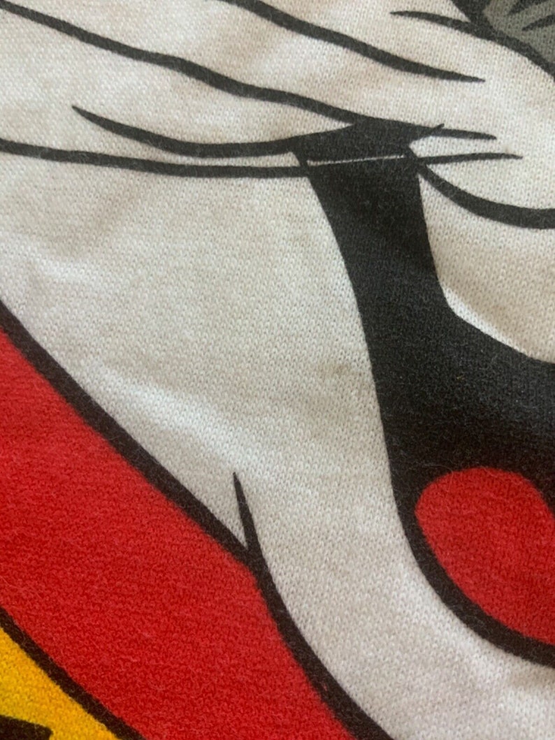 May include: Close-up of a white, red, black, and yellow graphic design. The design features a cartoon character's face, with a white face, black eyes and ears, and a red mouth. The background is a textured fabric.