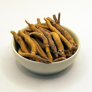 May include: A white ceramic bowl filled with dried, tan-colored roots. The roots are long, curved, and have a rough texture. The bowl is set against a plain white background, highlighting the natural color and form of the roots.