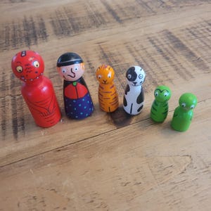 Può includere: Un set di sette bambole di legno con perni, dipinte in vari colori e design. Le bambole includono un drago rosso, una donna con un cappello nero e un vestito a pois, una tigre arancione, un cane bianco e nero e due rane verdi.