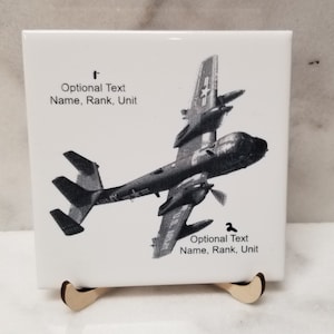 May include: A white ceramic tile with a black and white image of a military aircraft. The text "Optional Text Name, Rank, Unit" appears twice on the tile.