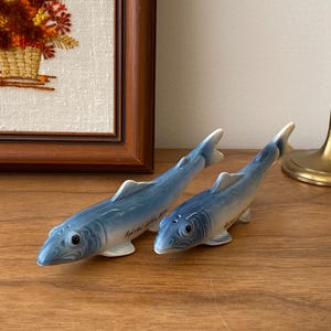 Puede incluir: Dos figuras de peces azules de cerámica con aletas blancas. Los peces tienen una textura y están colocados sobre una superficie de madera. El texto "Sylvia Lake 1974" está escrito en el lateral de un pez. En el fondo, un bordado enmarcado de flores.