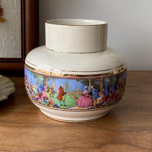 May include: Cream ceramic vase with a gold rim and a detailed, colourful landscape scene around the base. The scene depicts figures in period clothing in a garden setting. The vase has a cylindrical top.