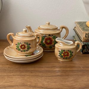 May include: A vintage tea set with a floral design. The set includes a teapot, creamer, sugar bowl, and saucers. The pieces are a light tan color with a red, yellow, and green floral pattern. The set is displayed on a wooden surface.