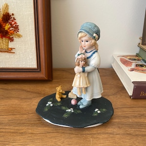 Könnte beinhalten: Porzellanfigur eines jungen Mädchens mit blauem Hut und Kleid, das eine Puppe hält. Die Figur steht auf einem schwarzen Sockel mit einem Teddybär und einem rosa Ball. Der Hintergrund umfasst eine gerahmte Blumenstickerei und einen Stapel Bücher.