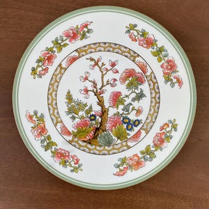 Może przedstawiać: Biały talerz ceramiczny z zielonym rantem zdobiony jest wzorem kwiatowym. Środek talerza ma geometryczny wzór w kolorze złotym i białym z kwiatowym wzorem w kolorze różowym i zielonym. Talerz ma delikatny wzór kwiatowy wokół krawędzi.