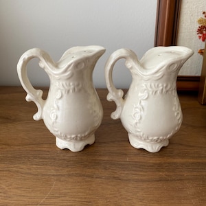 May include: A pair of off-white ceramic salt and pepper shakers, each shaped like a small jug. They feature a handle and floral relief detailing. The shakers are placed on a wooden surface.