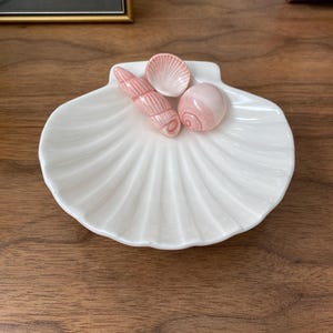 Può includere: Un piatto bianco in ceramica a forma di conchiglia con tre conchiglie rosa in ceramica all'interno. Le conchiglie sono una conchiglia a spirale, una capasanta e una conchiglia rotonda. Il piatto ha una texture a coste.