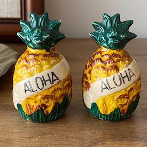 May include: A pair of pineapple-shaped salt and pepper shakers. The shakers are yellow, brown, and green with the word "ALOHA" printed on a white banner. The ceramic shakers have a textured surface.