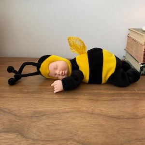 Peut inclure: Une poupée au teint pâle et aux yeux fermés, vêtue d'un costume de bourdon en peluche douce. La tenue présente un corps rayé jaune et noir, une capuche jaune avec des antennes noires et des ailes texturées jaunes et transparentes au dos.